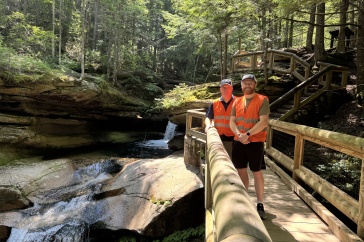 Two men wearing blaze orange vests stand on a log bridge above a stream