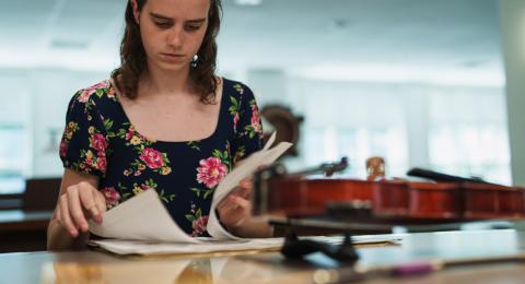 Woman looks though papers at a desk. A fiddle sits next to her.