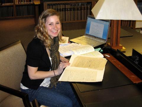 Stevens sits in an archive room at a desk with documents in front of her and a laptop on the desk as well. She is turning and smiling at the camera. 