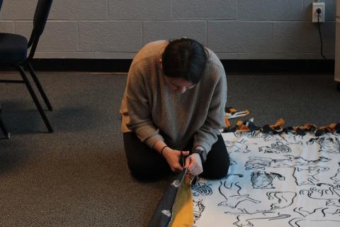 Student kneeling on floor cutting fleece fabric piece.