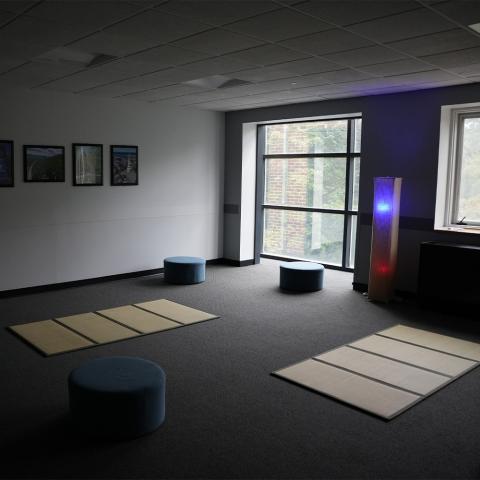 Room with mats on floor and round blue cusions, framed photos on wall, and a tall lamp with red and blue lights near large windows.