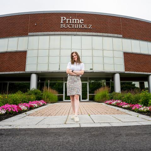 Jill standing with her arms crossed and smiling in front of the Prime Buccholz building. There are flowers aligning the walkway to the door in the background. 