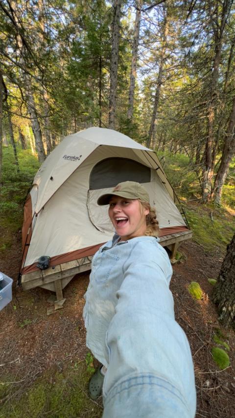 Briella smiling with a tent and the forest in the background. 