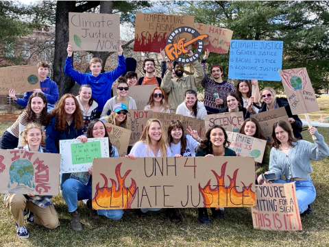 Students smiling holding posters with slogans for a Climate Justice Rally. The main poster says "UNH 4 Climate Justice"