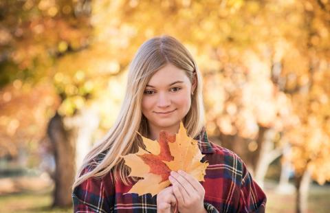 Courtney, a woman with long blonde hair, stands in a red flannel shirt among autumn leaves holding a fan of leaves towards the camera
