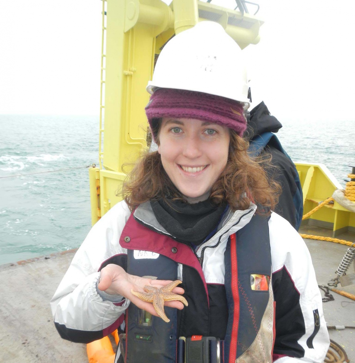 Woman on research vessel wears a hardhat and smiles while holding a seastar.