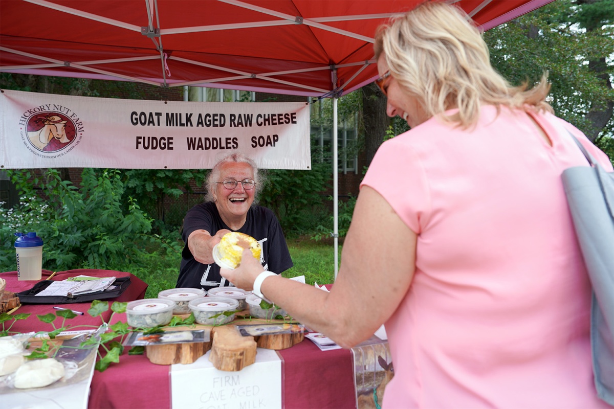 A farmers’ market vendor who selling goat cheese, fudge, soap and waddles hands a product to a customer.