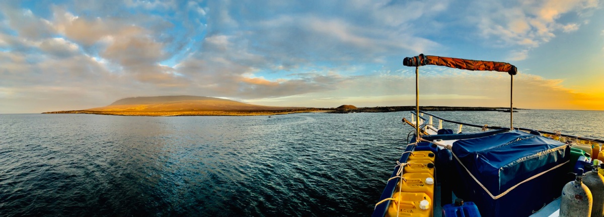 In the foreground is a boat in the water and in the background is an island rising up out of the water. The sky is blue and partly cloudy.