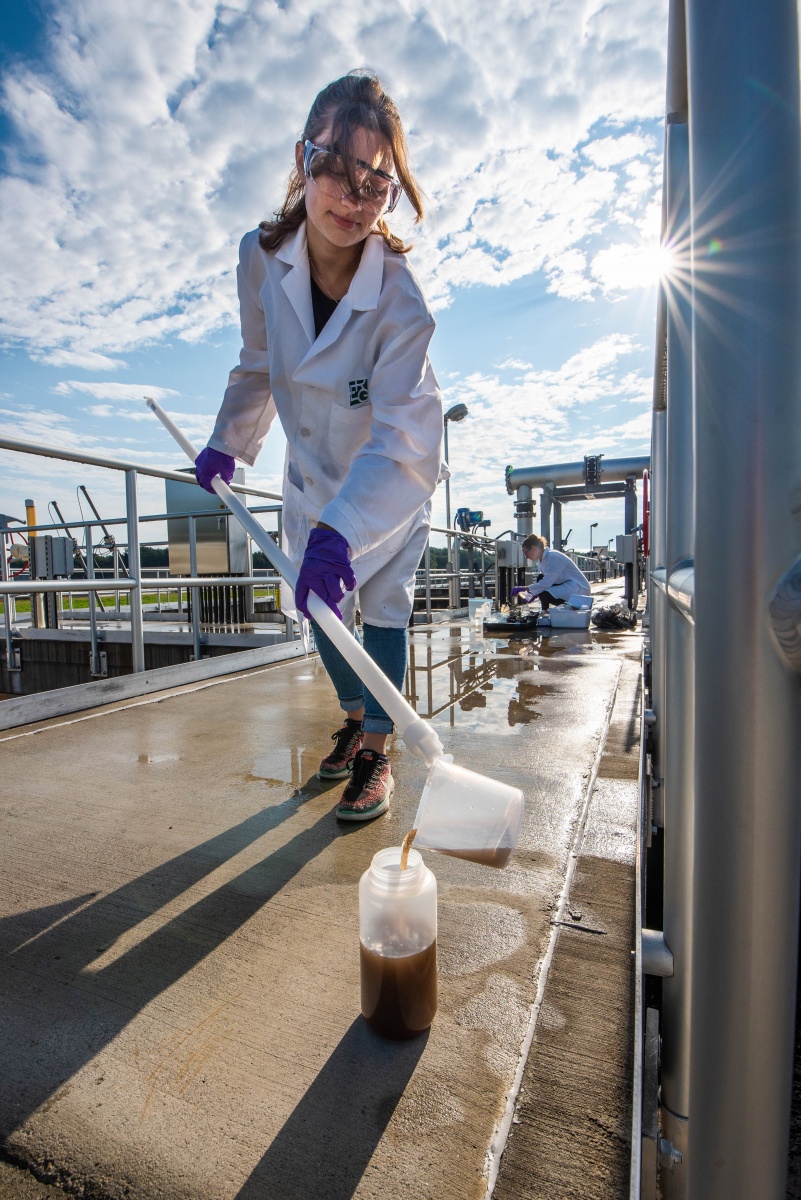 Woman wearing white lab coat samples wastewater outside with bright sun in background