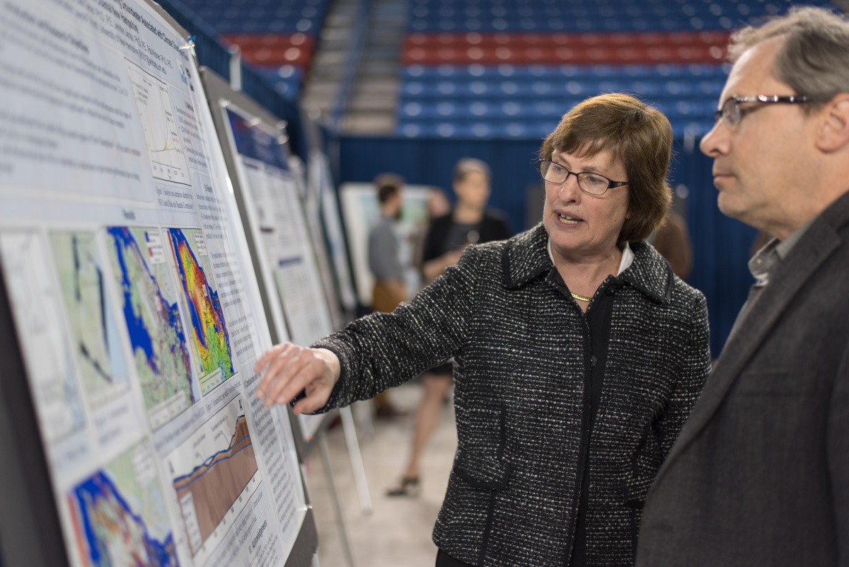 Woman in a suit explains a scientific poster to an onlooker