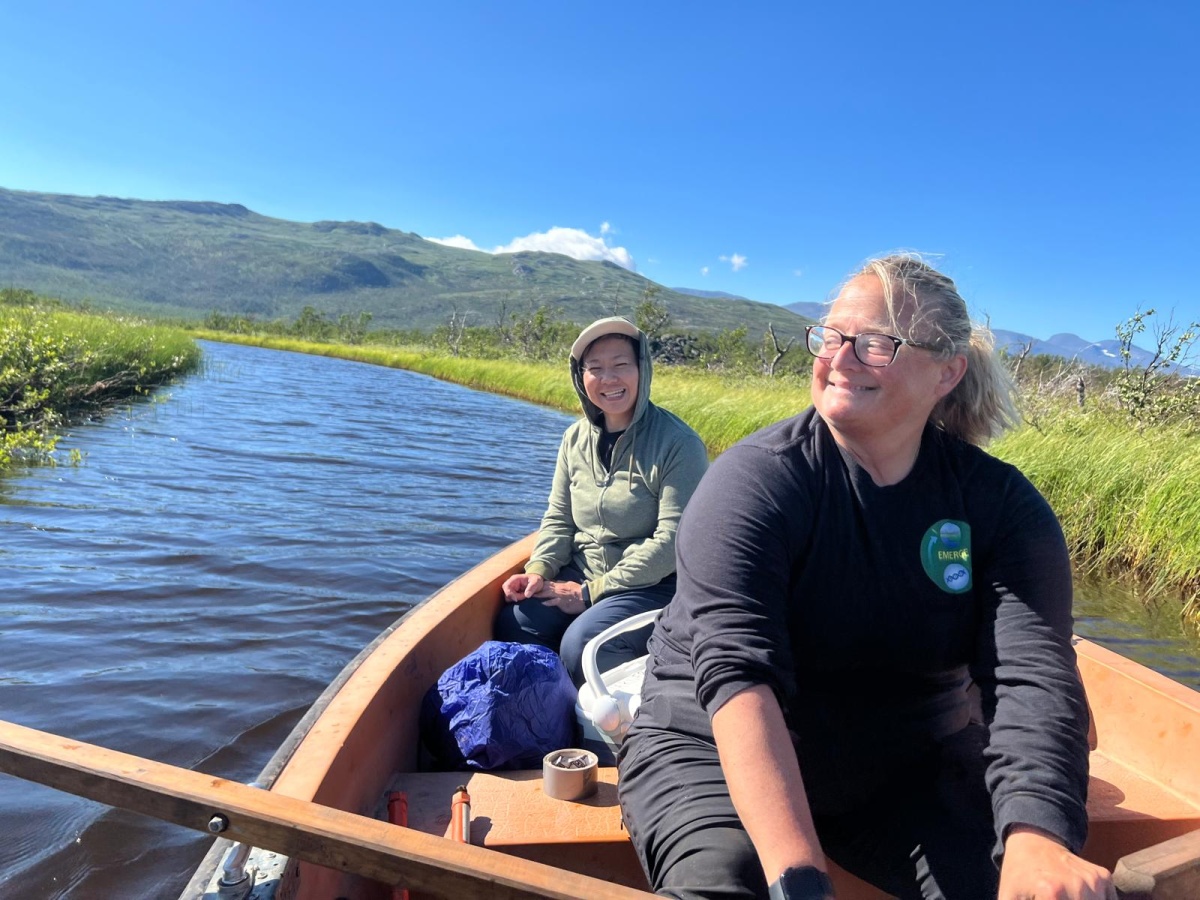 Two women row a wooden boat beneath snow-capped mountains.