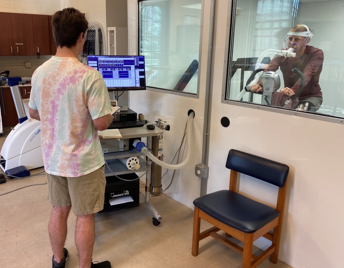 Researcher looks at a computer while a subject rides an exercise bicycle behind glass