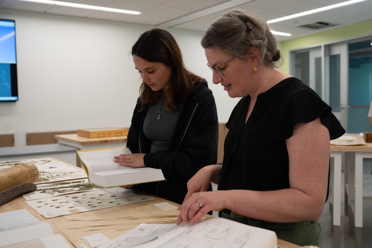 A photo showing two women standing at a table and looking through specimens from the Hodgdon Herbarium of an extinct crabgrass found only in New Hampshire at one time.