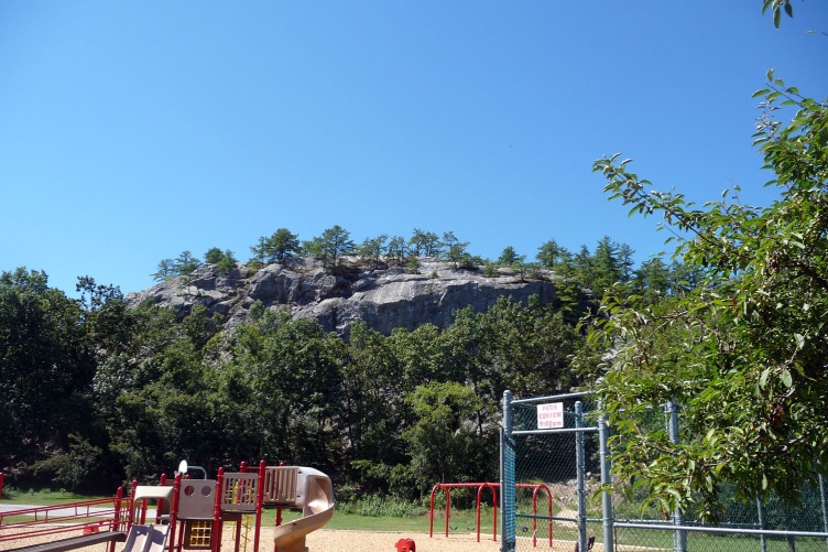 A photo of a prominent rock formation in the background and, in the foreground, a playground next to that rock. The rock is Rock Rimmon in Manchester, NH.