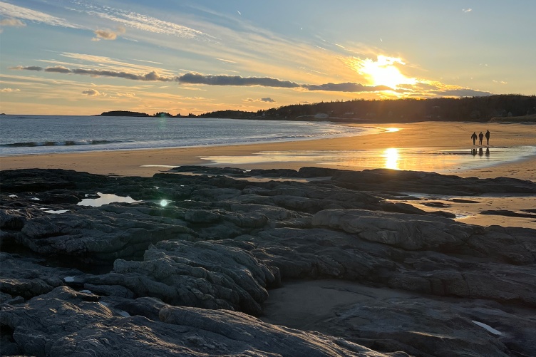 A scenic photo of the Gulf of Maine shoreline showing two people walking into a sunset.