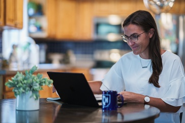 Woman working on computer at a kitchen table with a UNH mug in the foreground