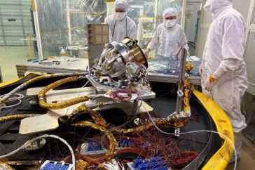 Three people wearing white Tyvek suits gather around a space instrument in a laboratory. 