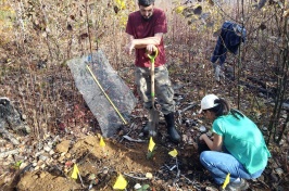 Two researchers (1 male standing, holding a shovel, 1 female squatting next to a whole) work in a prescribed burn site to plant potted red oak trees.
