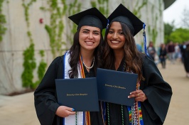 Two female graduates pose with their diplomas in their caps and gowns outside of commencement