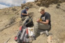 Two male paleontologists collect rocks on a dusty hillside