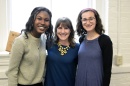 A photo of three woman, 1 with dark skin and 2 with light skin. The person in the middle has her arms around the other 2 people.