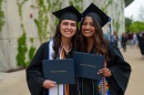 Two female graduates pose with their diplomas in their caps and gowns outside of commencement