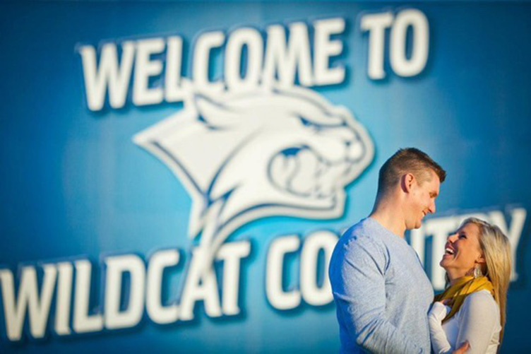 Man and woman look into each other's eyes in front of a stadium sign that reads Welcome to Wildcat Country