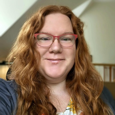 Kate Filanoski is a white woman with long wavy red hair. She is smiling at the camera and wearing red rimmed glasses and a dark gray blazer.